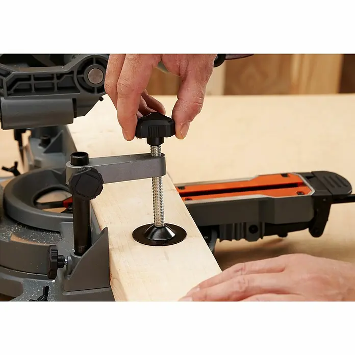 Black+Decker Sierra ingletadora BES702 - 1.500 W, Diámetro hoja de sierra: 216 mm, Número de revoluciones de marcha en vacío: 5.000 r.p.m. - Primer plano de una sierra de inglete con una mano apretando una abrazadera sobre un trozo de madera.