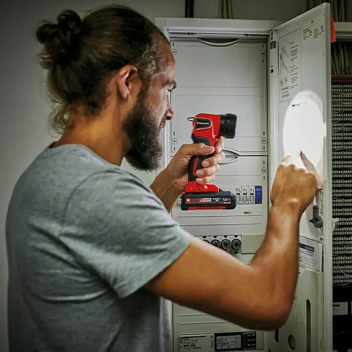 Einhell Power X-Change 18V Akku-Lampe TC-CL 18 Li H-Solo - 18 V, Ohne Akku, Lichtstrom: 280 lm - Ein Mann beleuchtet mit einer Einhell Akku-Lampe einen Verteilerkasten. Die Lampe ist rot und schwarz und wird mit einem Akku betrieben.