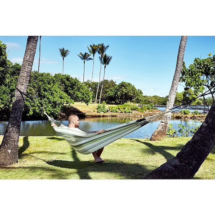 La Siesta Hängematte Brisa - L x B: 350 x 160 cm, Cedar - Ein Mann entspannt in einer gestreiften Hängematte zwischen zwei Palmen, die perfekte Entspannung im Freien.