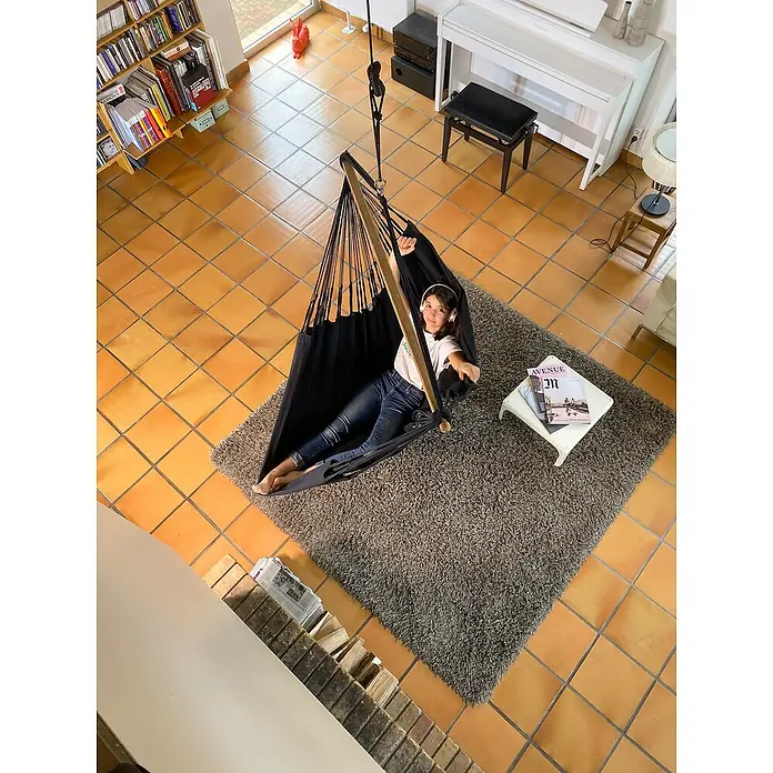 La Siesta Hängesessel Habana Comfort - L x B x H: 180 x 115 x 160 cm, Onyx - Eine junge Frau entspannt in einer schwarzen Hängematte mit Holzrahmen, Kopfhörer auf, auf einem grauen Teppich in einem gemütlichen Wohnzimmer.