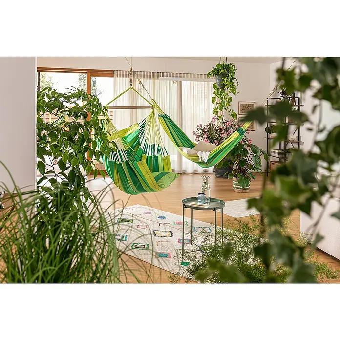 La Siesta Hängematte Modesta - L x B: 350 x 160 cm, Jungle, Double - Helle Inneneinrichtung mit grünen Hänge-Hängematten, Pflanzen und einem kleinen Tisch auf einem gemusterten Teppich für entspannte Momente.