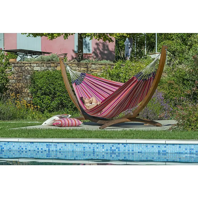 La Siesta Hängematte Modesta - L x B: 350 x 160 cm, Flamingo, Double - Eine Frau entspannt sich in einer farbenfrohen Hängematte mit Holzgestell neben einem Pool, umgeben von üppigem Grün.