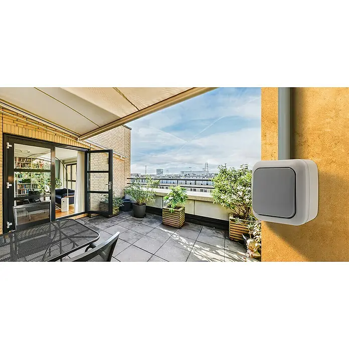 Siemens Delta IP54 Feuchtraumtaster SIE028653 - Grau, Aufputzmontage, IP54 - Nahaufnahme eines grauen Aufputz-Lichtschalters an einer gelben Wand auf einer Terrasse mit Blick auf die Stadt.