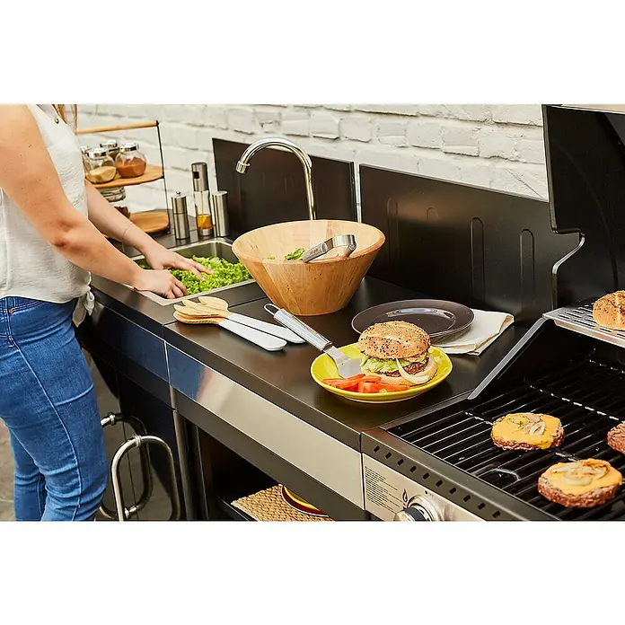 Mueble auxiliar modular para barbacoa Fidgi - Negro/Plateado - Escena de una persona lavando ensalada y preparando una hamburguesa con foco en la preparación de alimentos al aire libre.