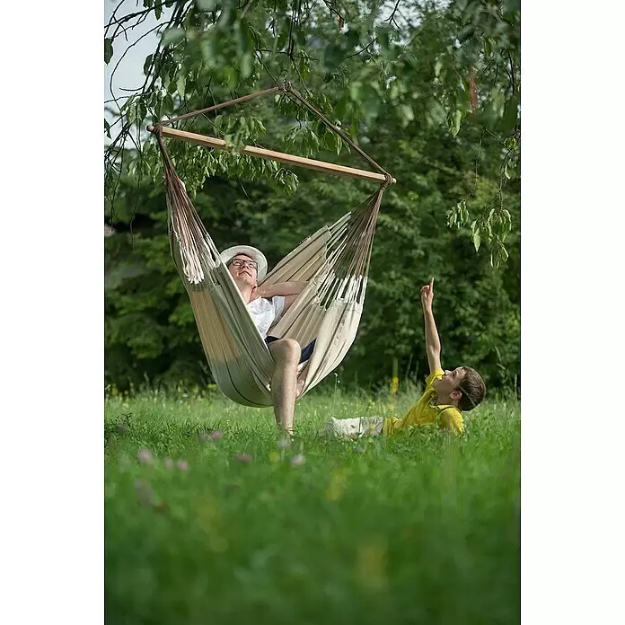 La Siesta Hängesessel Habana Kingsize - L x B x H: 210 x 130 x 170 cm, Nougat - Ein Mann entspannt in einem beigen Hängesessel mit Holzspreizstab, der an einem Baum hängt. Ein Kind liegt im Gras und zeigt nach oben.
