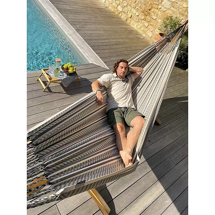 La Siesta Doppelhängematte Flora - L x B: 400 x 180 cm, Zebra, Ausführungseigenschaft: Kingsize - Ein Mann entspannt sich in einer bequemen, gestreiften Stoffhängematte auf einer Holzterrasse neben einem Pool.