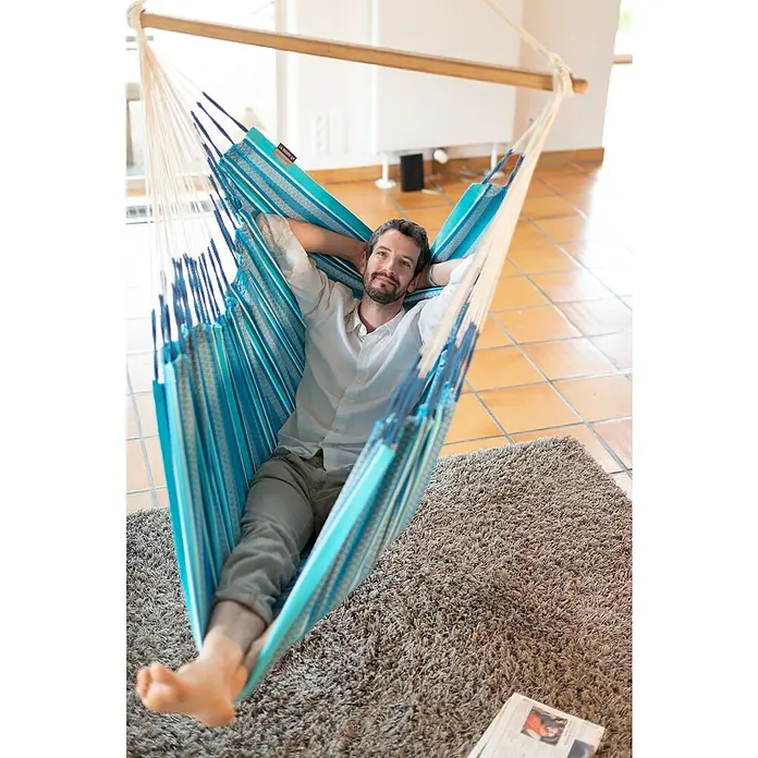 La Siesta Hängesessel Habana Kingsize - L x B x H: 210 x 130 x 170 cm, Azure - Ein Mann entspannt in einer blauen Hängematte mit Holzspreizstab auf einem beigen Teppich. Die Hängematte lädt zum Verweilen und Entspannen ein.