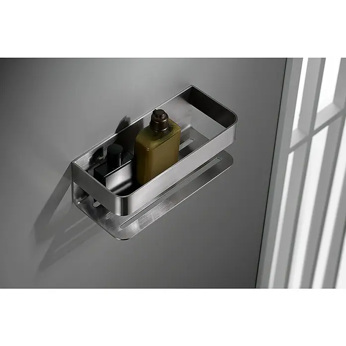 Imex Cesta de baño Mayura - 11 x 25,6 x 8 cm, Acero inoxidable, Níquel - Una estantería de ducha plateada montada en una pared gris, con una botella de gel de ducha y una botella pequeña dentro, junto a una ventana con persianas.