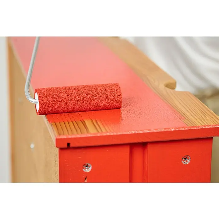 swingcolor Buntlack Acryl - Feuerrot, Glänzend, 750 ml, Wasserbasiert - Ein Farbroller trägt rote Farbe auf eine Holzoberfläche auf, wodurch ein glänzendes, frisch gestrichenes Finish entsteht.