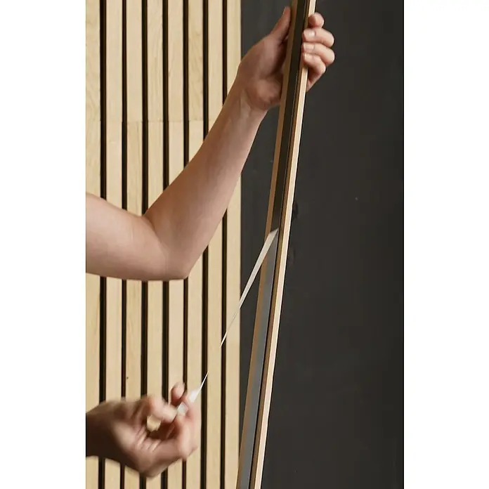 b!design Wandpaneel Wall Smart - Eiche Hellbraun, 2.500 x 35 x 9 mm - Eine Person entfernt die Schutzfolie von einem Akustikpaneel mit Holzrahmen, der an einer Wand befestigt ist. Das Paneel dient der Schallabsorption.