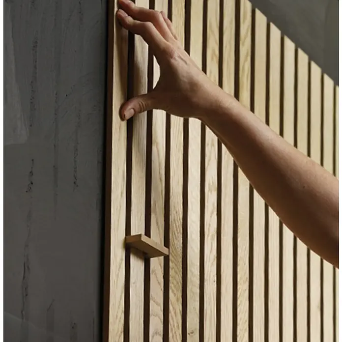 b!design Wandpaneel Wall Smart - Eiche Hellbraun, 2.500 x 35 x 9 mm - Nahaufnahme einer Holzlamellenwand, die an einer grauen Betonwand befestigt ist, wobei eine Hand die Textur und das Design betont.