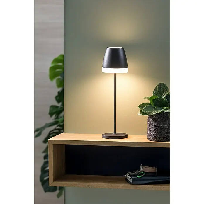 Lavida Akku-Tischleuchte Loria - 2 W, Schwarz, Warmweiß - Elegante schwarze Tischleuchte auf einem Holzregal, die ein warmes Licht ausstrahlt, neben einer Topfpflanze und persönlichen Gegenständen.