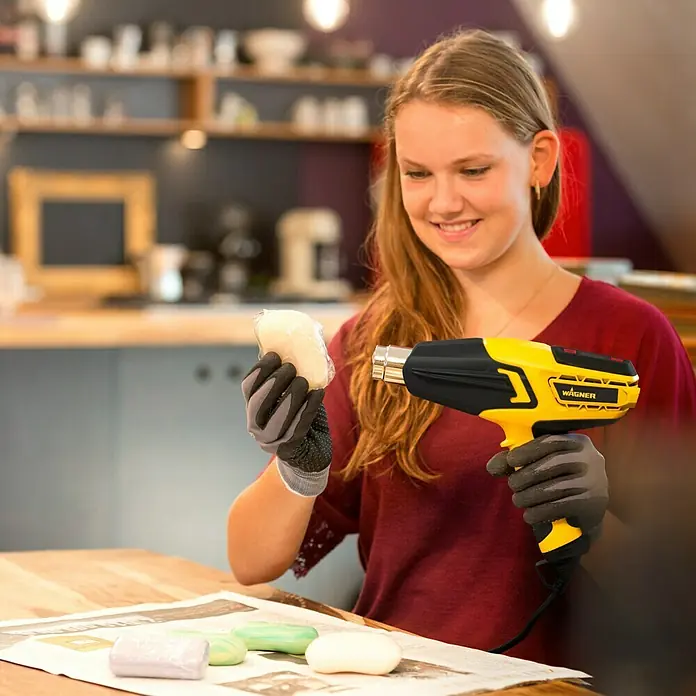 Wagner Heißluftgebläse Furno 750 - 2.000 W - Frau, die ein Wagner-Heißluftgebläse benutzt, um Seife zu verpacken. Das Heißluftgebläse ist gelb und schwarz, und die Seife ist in Plastik verpackt.