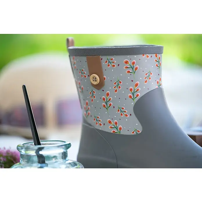 Black Fox Gummistiefel Bella - Schuhgröße: 38, Grau - Nahaufnahme eines grauen Gummistiefels mit floralem Muster, verziert mit einem Knopf und einer Schlaufe, vor einem verschwommenen, natürlichen Hintergrund.