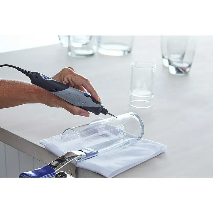 Dremel Elektro-Multifunktionswerkzeug 2050 Stylo+ - 9 W - Detailaufnahme einer Hand, die mit einem Dremel-Werkzeug ein Glas mit filigranen Mustern verziert, während es mit einer Klemme fixiert wird.