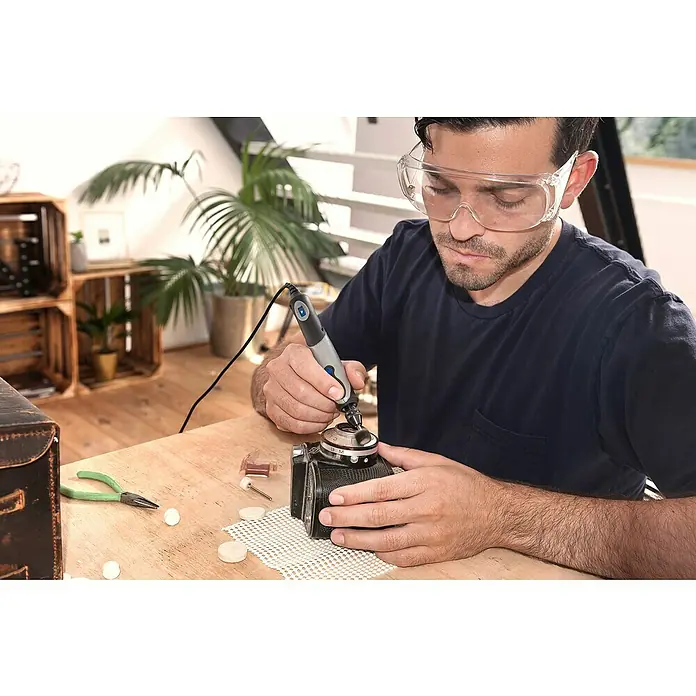 Dremel Elektro-Multifunktionswerkzeug 2050 Stylo+ - 9 W - Ein Mann, der eine Dremel-Rotationswerkzeug zum Reinigen einer Vintage-Kamera verwendet, mit Zubehör und Schutzbrille auf einem Holztisch.
