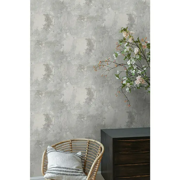 Papel pintado Cemento Liso - Gris, Efecto cemento, 10 x 0,53 m - Un primer plano de una cómoda con decoración de ramas en flor, una silla de ratán con cojín y una pared gris con textura.