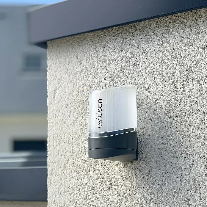 Avidsen Luces intermitentes LED para mecanismos de puerta de garaje - 8 W - Primer plano de una luz exterior Avidsen con un diseño limpio y moderno y una carcasa robusta montada en una pared texturizada.