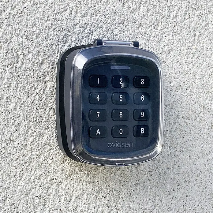 Avidsen Teclado de código inalámbrico - Alcance: 80 m - Primer plano de una caja fuerte con teclado Avidsen montada en una pared. El dispositivo cuenta con una cubierta transparente y botones numerados del 1 al 9, A, 0 y B.