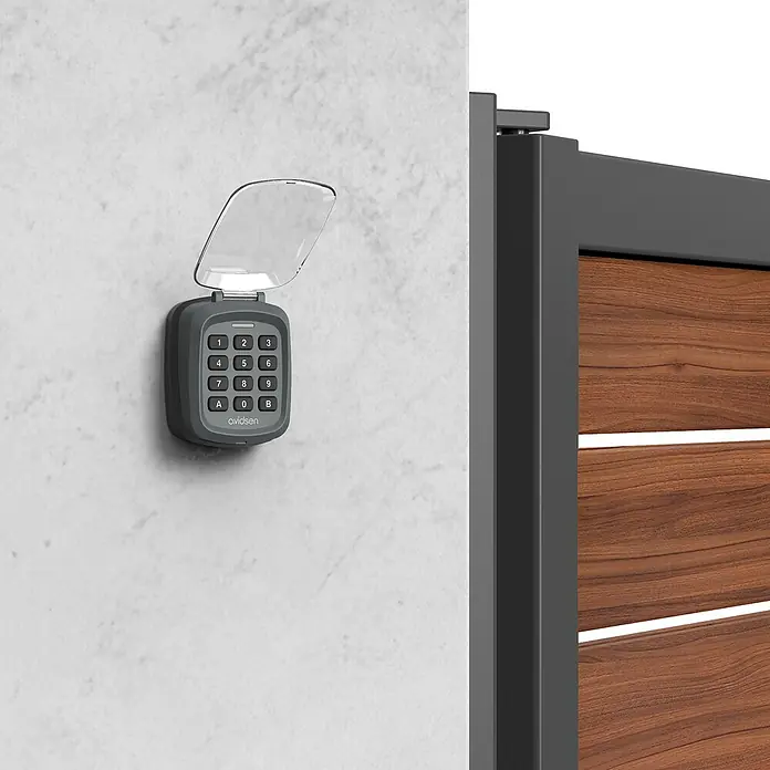 Avidsen Teclado de código inalámbrico - Alcance: 80 m - Teclado Avidsen con tapa abatible para control de acceso seguro, montado en una pared blanca junto a una puerta de madera moderna.