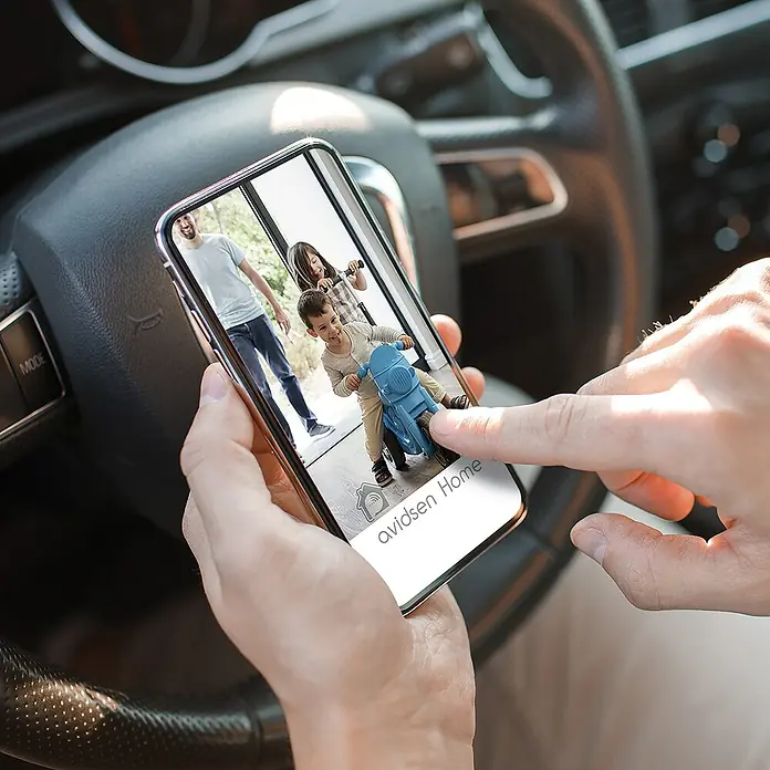 Avidsen Módulo de comando inalámbrico WiFi - 50 m - Primer plano de un smartphone que muestra la aplicación Avidsen Home, sostenido frente a un volante, mostrando un video en vivo de niños jugando.