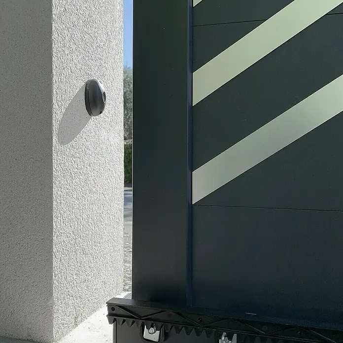 Avidsen Mecanismo de puerta de garaje Orane 510 con brazo articulado - Anchura puerta: 0 m - 2,5 m - Una puerta moderna con rayas diagonales se encuentra junto a una pared blanca con un sensor redondo. La puerta es oscura y minimalista.