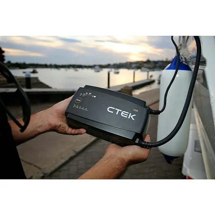CTEK Automatik-Ladegerät M 25 EU - Kapazität: 40 Ah - 500 Ah - Ein CTEK M25 Batterieladegerät für 12V Batterien wird vor einer Küstenlandschaft gehalten, ideal für Boote und Fahrzeuge.
