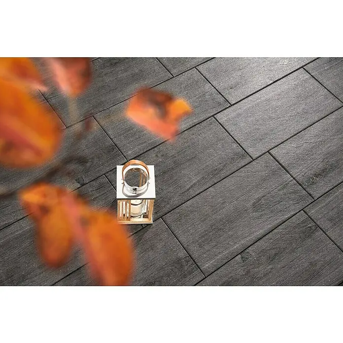 Diephaus Terrassenplatte Nashville - 60 x 40 x 4 cm, Graphit, Beton - Stilvolles Foto von Bodenfliesen in Holzoptik mit einer dekorativen Laterne und herbstlichen Blättern im Vordergrund, ideal für Wohnraumgestaltung.