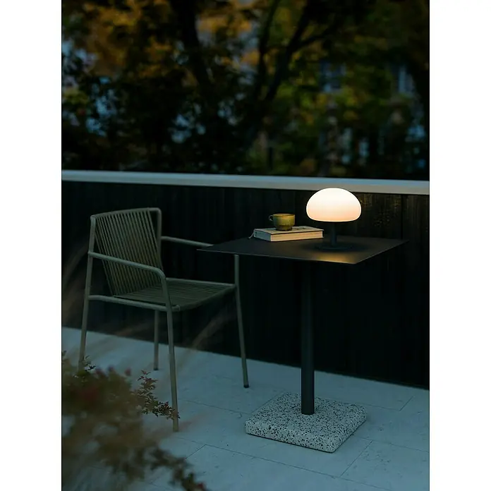 Nordlux Sponge Akku-Tischleuchte - Schwarz, Weiß, Warmweiß, 4,8 W, IP65 - Eine moderne Tischleuchte mit Pilzdesign beleuchtet einen kleinen Tisch auf einer Terrasse, neben einem passenden Stuhl.