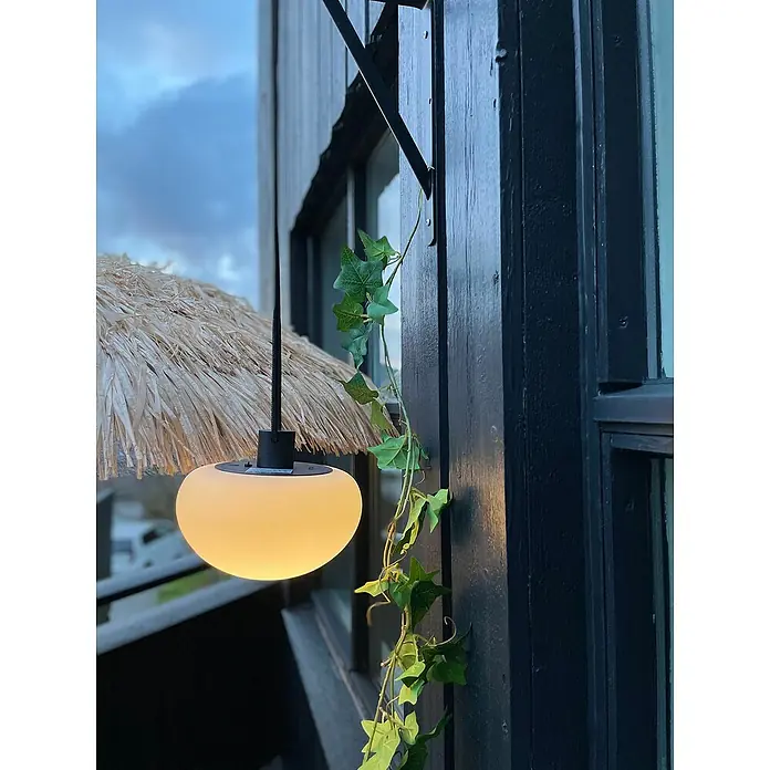Nordlux Sponge Akku-Pendelleuchte Sponge - 4,8 W, Warmweiß - Dekorative Außenleuchte mit einem kugelförmigen Lampenschirm und Strohschirm, aufgehängt an einer dunklen Hauswand mit Efeu.