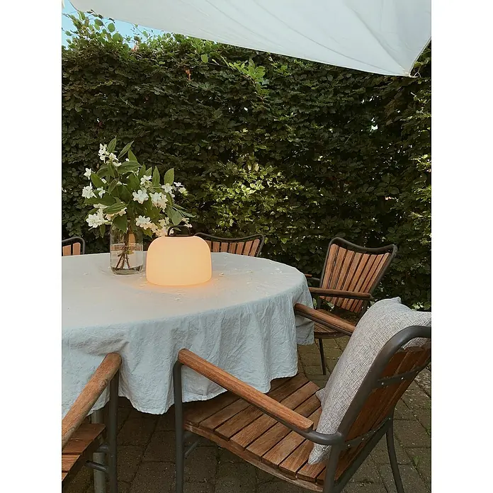 Nordlux Akku-Pendelleuchte Kettle 22 - 4,8 W, Warmweiß - Ein Gartenbereich mit einem runden Tisch, bedeckt mit einem weißen Tuch, einer Lampe und Stühlen mit Holzsitzen. Die Lampe ist der Fokus.