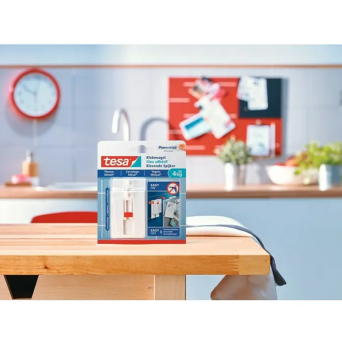 Tesa Clavo adhesivo ajustable - Blanco, Carga soportada: 4 kg, 2 ud. - Clavos adhesivos Tesa para azulejos y metal, reposicionables y reutilizables, expuestos sobre una mesa de madera con fondo de cocina.