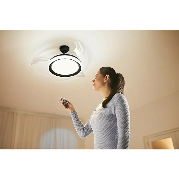 Philips Ventilador de techo Bliss Negro - Transparente, 51 cm, 28 W - 35 W, Blanco cálido - Una mujer opera una lámpara de techo con un ventilador integrado mediante un mando a distancia, combinando iluminación y circulación de aire.