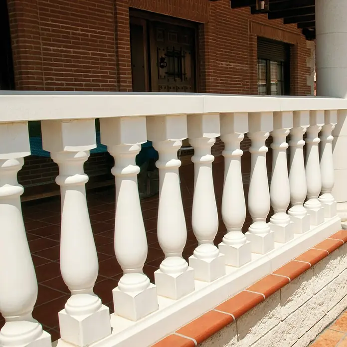 Balaustrada Italia 75 - 12 x 12 x 75 cm, Blanco, Hormigón - Primer plano de una balaustrada blanca con pilares frente a una casa de ladrillo rojo, destacando la elegancia arquitectónica y la artesanía.
