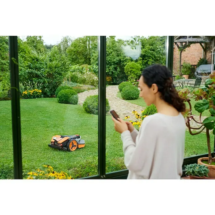 Worx PowerShare 20V Mähroboter Landroid Vision L1300 - 1.300 m², Schnittbreite: 22 cm, Kamera-gestützte Navigation - Frau steuert einen Gardena Mähroboter mit ihrem Smartphone aus einem Wintergarten, ein gepflegter Garten im Hintergrund.