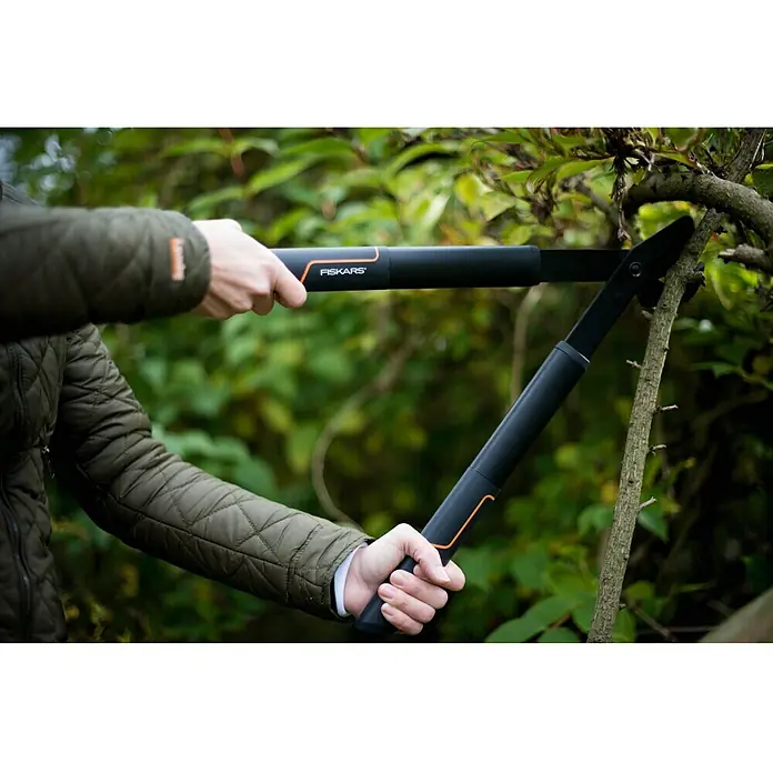 Fiskars SingleStep Hand-Astschere L28 - Länge: 57 cm, Max. Aststärke: 35 mm, Stahl, Schwarz/Orange - Eine Person verwendet eine Fiskars Astschere mit Teleskopstiel, um einen Ast zu schneiden. Die Astschere ist schwarz mit orangen Akzenten.