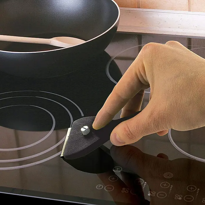 Rascador para vitrocerámica - Largo: 13,9 cm, Específico para: Placas de cocina - Un rascador de vidrio limpia una placa vitrocerámica, mientras que una olla con cuchara de madera está al fondo. ¡Ideal para la cocina!