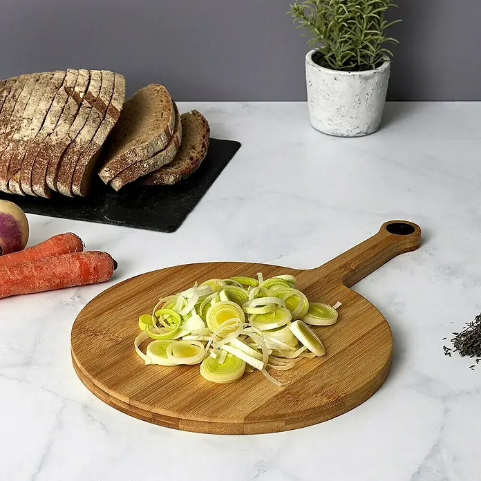 Tabla de cortar Redonda - Ø x L: 24 x 37,5 cm, Bambú - Una tabla de cortar redonda de madera con puerros en rodajas y un mango, ideal para servir o como tabla de cortar de cocina.