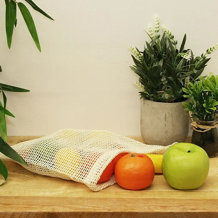 Bolsa de malla lavable - 2 ud., An x Al: 30 x 38 cm - Bolsa de compra reutilizable llena de fruta fresca como manzanas y naranjas sobre una mesa de madera, junto a plantas.