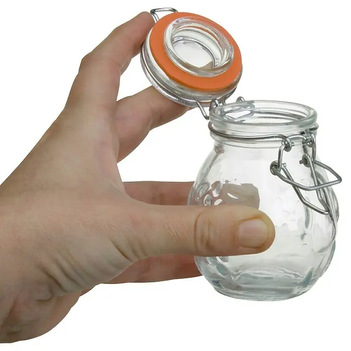 Frasco con cierre mecánico - Vidrio, Surtido - Un frasco de conserva con cierre de alambre y junta naranja sostenido abierto por una mano, ideal para conservas, con diseños de uvas en relieve.