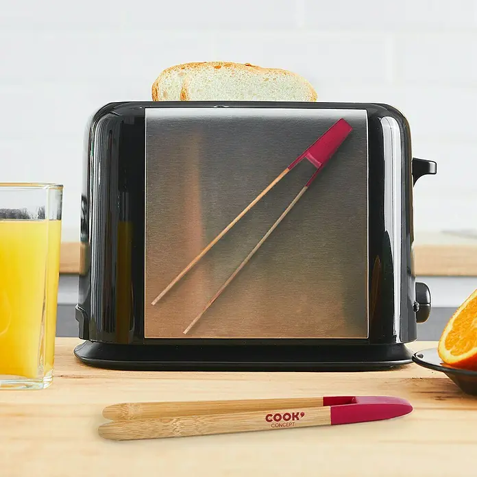 Pinza con imán - Largo: 240 mm, Bambú - Primer plano de una tostadora con una rebanada de pan tostado y unas pinzas con mangos rojos delante de una mesa de desayuno.