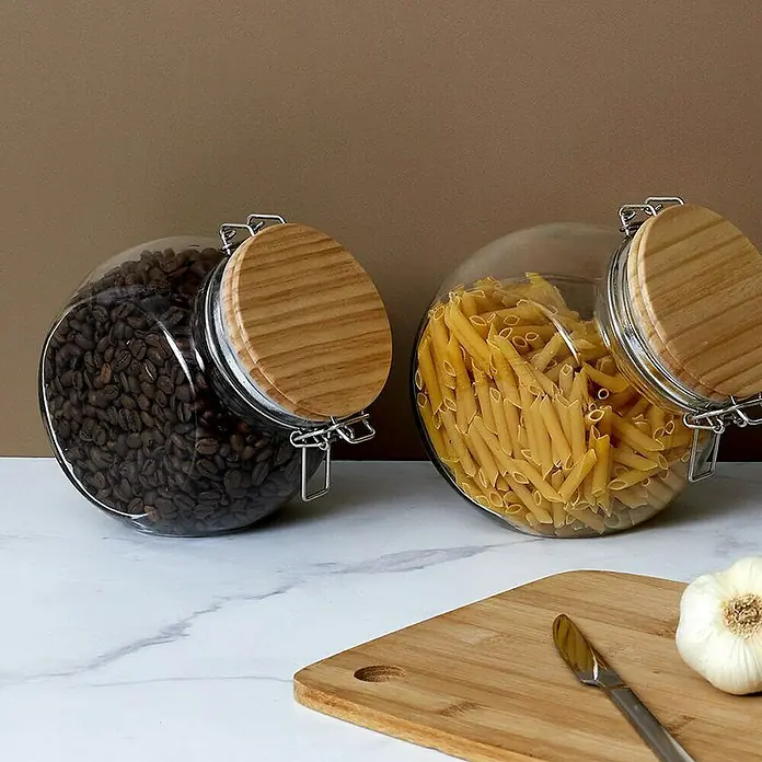 Frasco con cierre mecánico - Vidrio, 1,7 l - Dos tarros de almacenamiento con tapas de madera, uno lleno de granos de café y el otro de pasta, sobre superficie de mármol.