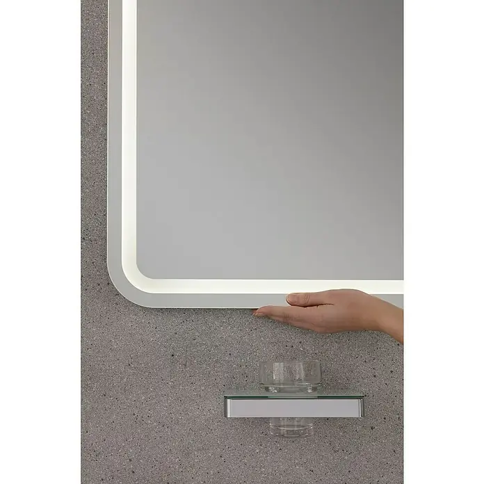 Lanzet Classico LED-Lichtspiegel - 100 x 70 cm - Das Bild zeigt einen beleuchteten Spiegel, der an einer Wand montiert ist, mit einer Hand, die ihn berührt, und einem kleinen Regal mit einem Glas darunter.