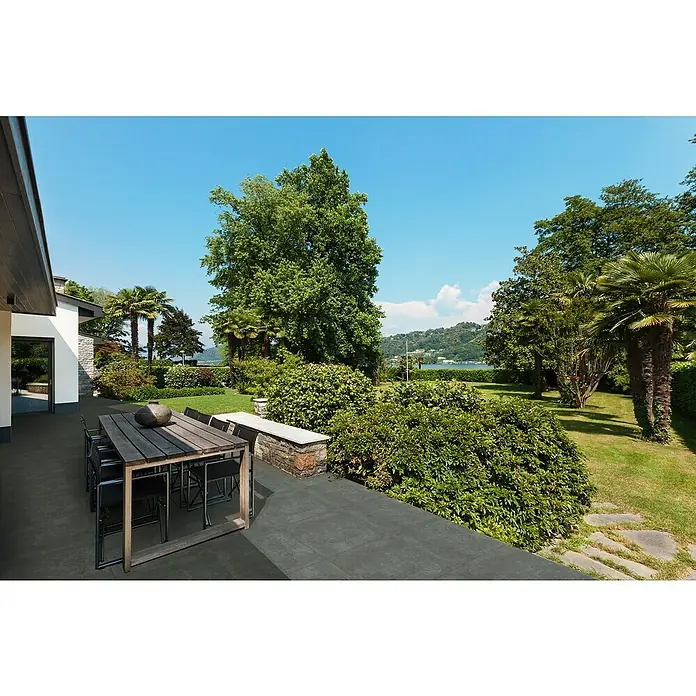 Terrassenfliese Manchester 2.0 - 60 x 60 x 2 cm, Schwarz, Matt - Im Freien steht ein Esstisch aus Holz mit schwarzen Stühlen auf einer Terrasse, umgeben von üppigem Grün und Bäumen.