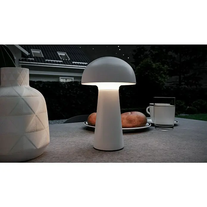 Trio Leuchten Akku-Tischleuchte Lennon - 2 W, Weiß, Warmweiß - Nahaufnahme einer modernen Pilzlampe mit warmem Licht, umgeben von stilvollen Objekten auf einem Tisch im Freien bei Nacht.