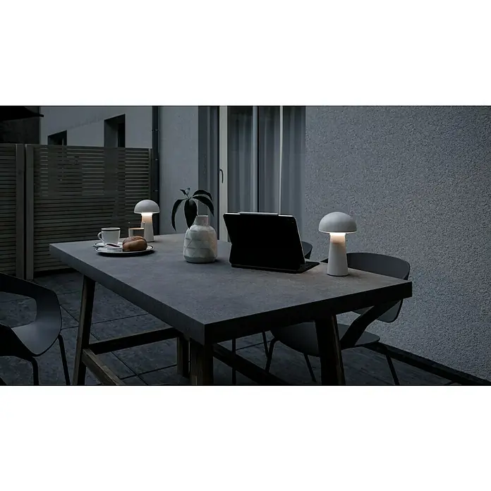 Trio Leuchten Akku-Tischleuchte Lennon - 2 W, Weiß, Warmweiß - Eine stimmungsvolle Außenaufnahme mit zwei leuchtenden Pilzleuchten auf einem Tisch mit Tablet, Gebäck und Pflanze.