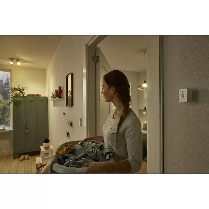 Philips Hue Bewegungsmelder - Weiß, L x B x H: 2 x 5,5 x 5,5 cm - Weißer Smart-Home-Bewegungssensor an einer Wand neben einer Tür, während eine Person mit einem Wäschekorb vorbeigeht.