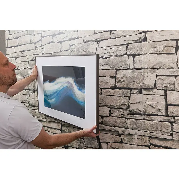Knauf Gipsplatten Metalldübel - Länge Dübel: 37 mm, Länge Schraube: 35 mm - Ein Mann hängt ein gerahmtes Kunstwerk mit abstraktem Design an einer Steinwand auf. Das Bild hat einen breiten, weißen Rand und einen dunklen Rahmen.