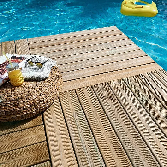 Cerland Loseta de madera Julia - Madera, 100 x 100 cm - Primer plano de una terraza de madera junto a la piscina con una bebida, un periódico y gafas de sol.