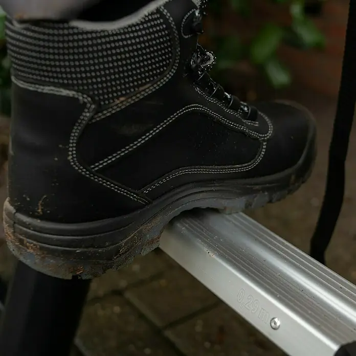Batavia Teleskopleiter Professional - B x H: 88 x 9 cm, Anzahl Tritte: 12 Stk., Arbeitshöhe: 4,6 m - Ein schwarzer, schmutziger Arbeitsstiefel steht auf einer silbernen Leitersprosse. Der Stiefel ist hoch geschnitten und hat Schnürsenkel.
